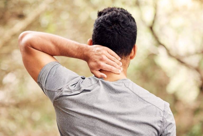 Head-Neck-Masses-evaluation Man in an outdoor setting holding the back of his neck with one hand. The image is from behind and shows the mans back and back of head.